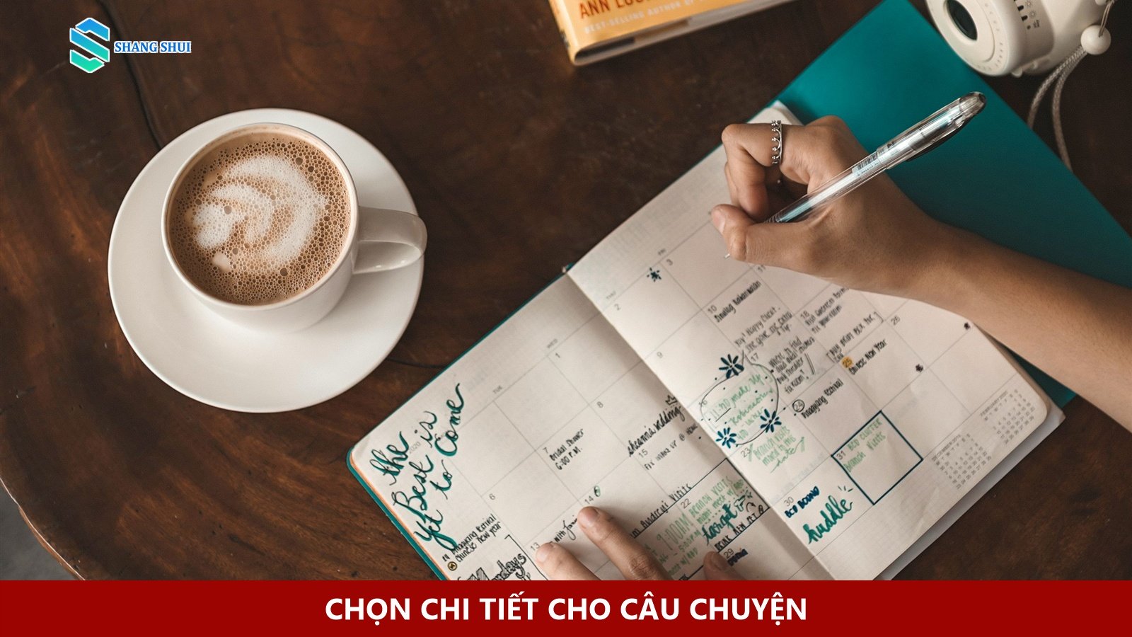 Bàn tay viết vào sổ cạnh ly cà phê, sách và máy ảnh.