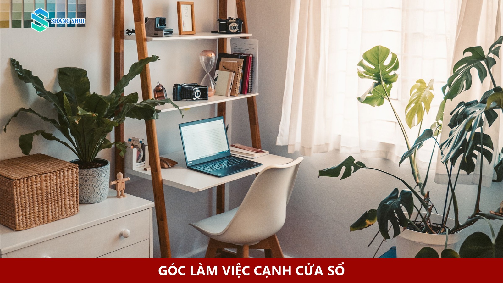 Bàn làm việc có laptop, ghế và cây xanh đặt cạnh cửa sổ có rèm mỏng.