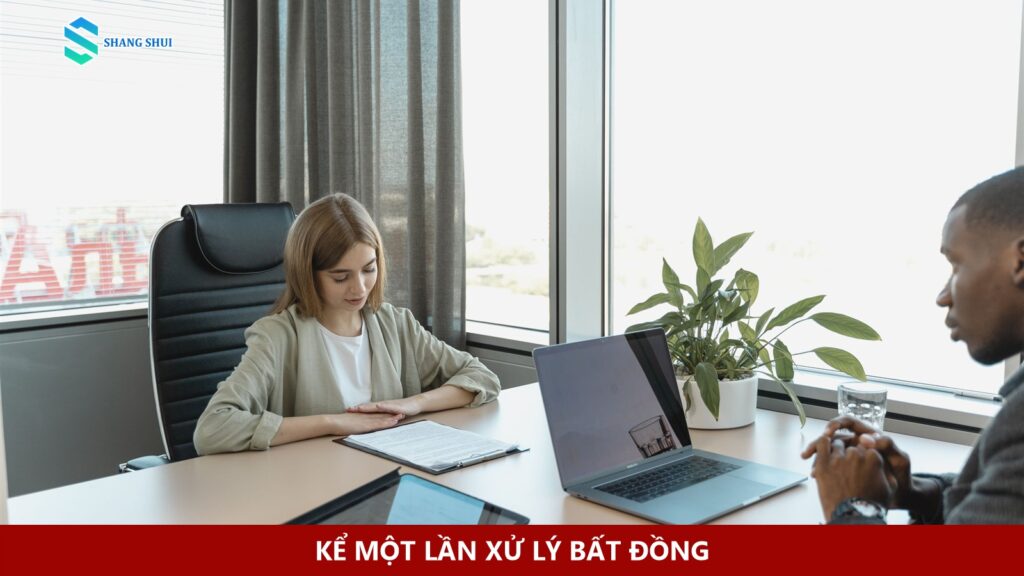 Hai người trao đổi qua bàn làm việc có laptop và tài liệu.