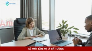Hai người trao đổi qua bàn làm việc có laptop và tài liệu.