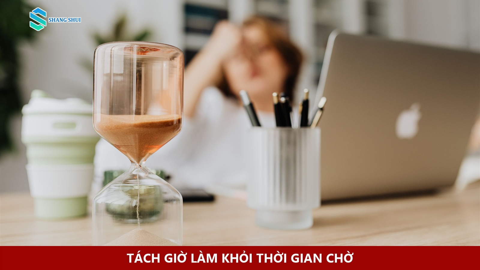Đồng hồ cát trên bàn, phía sau là laptop và một người ngồi làm việc.